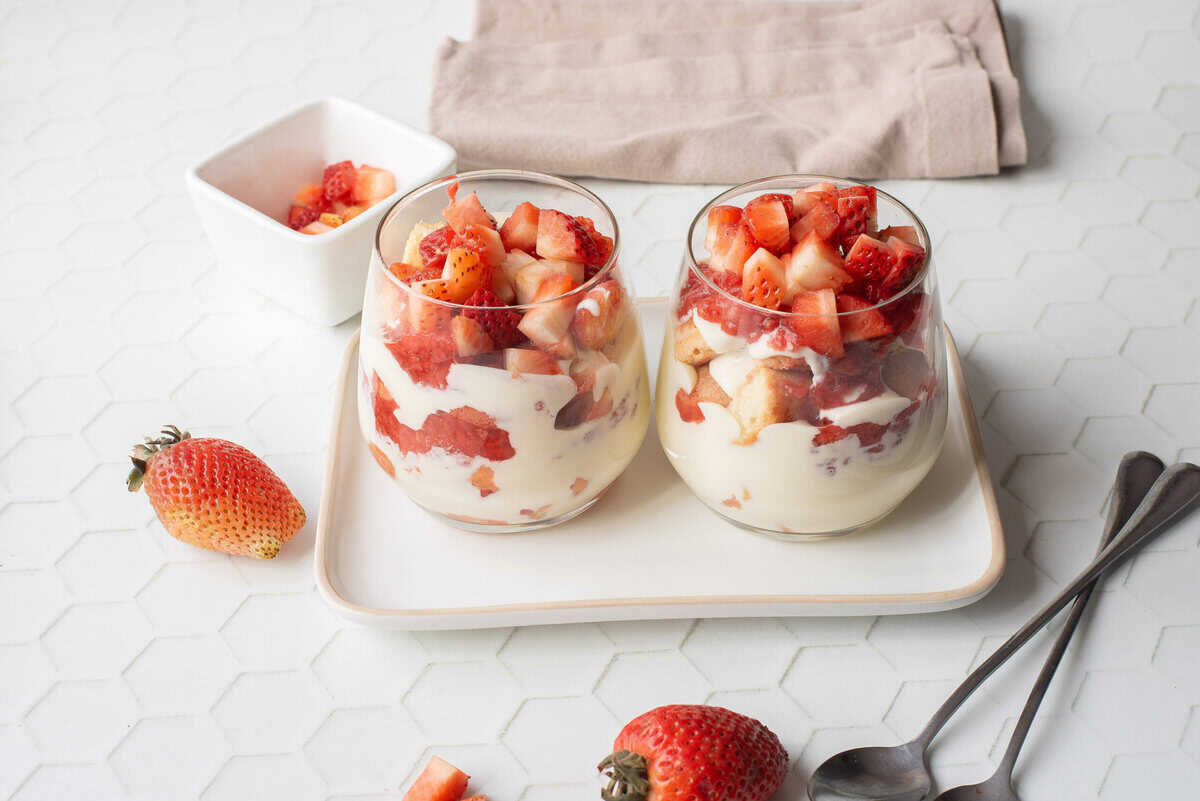 Two glass cups filled with layers of cream and chopped strawberries are placed on a white tray, with fresh strawberries and spoons nearby.