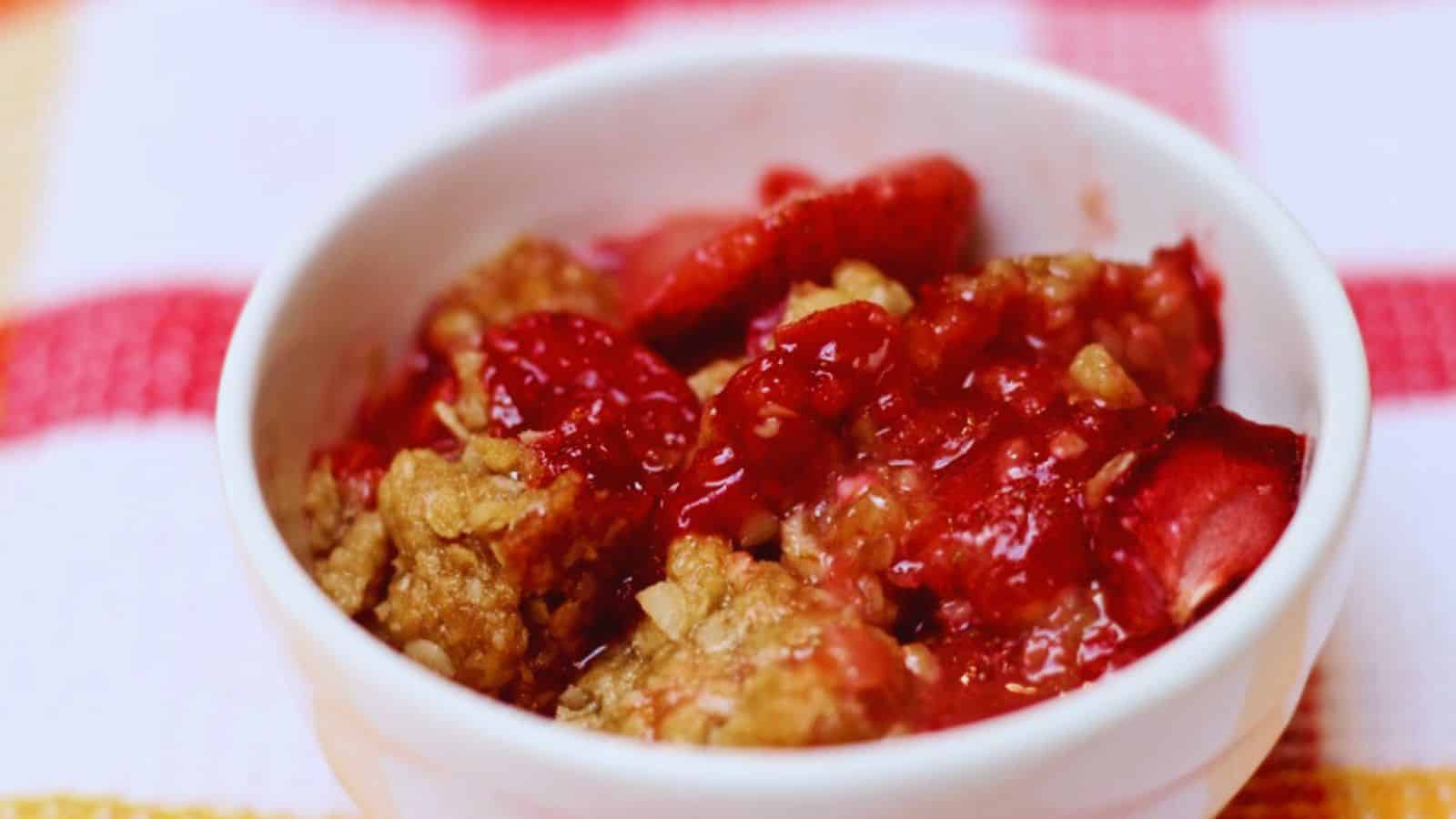 A white bowl filled with a baked dessert featuring strawberries and a crumbly oat topping, placed on a red and white checkered cloth.
