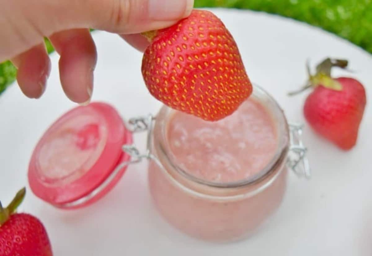 A hand holds a fresh strawberry above a glass jar filled with pink strawberry cream, with two more strawberries nearby on a white surface.