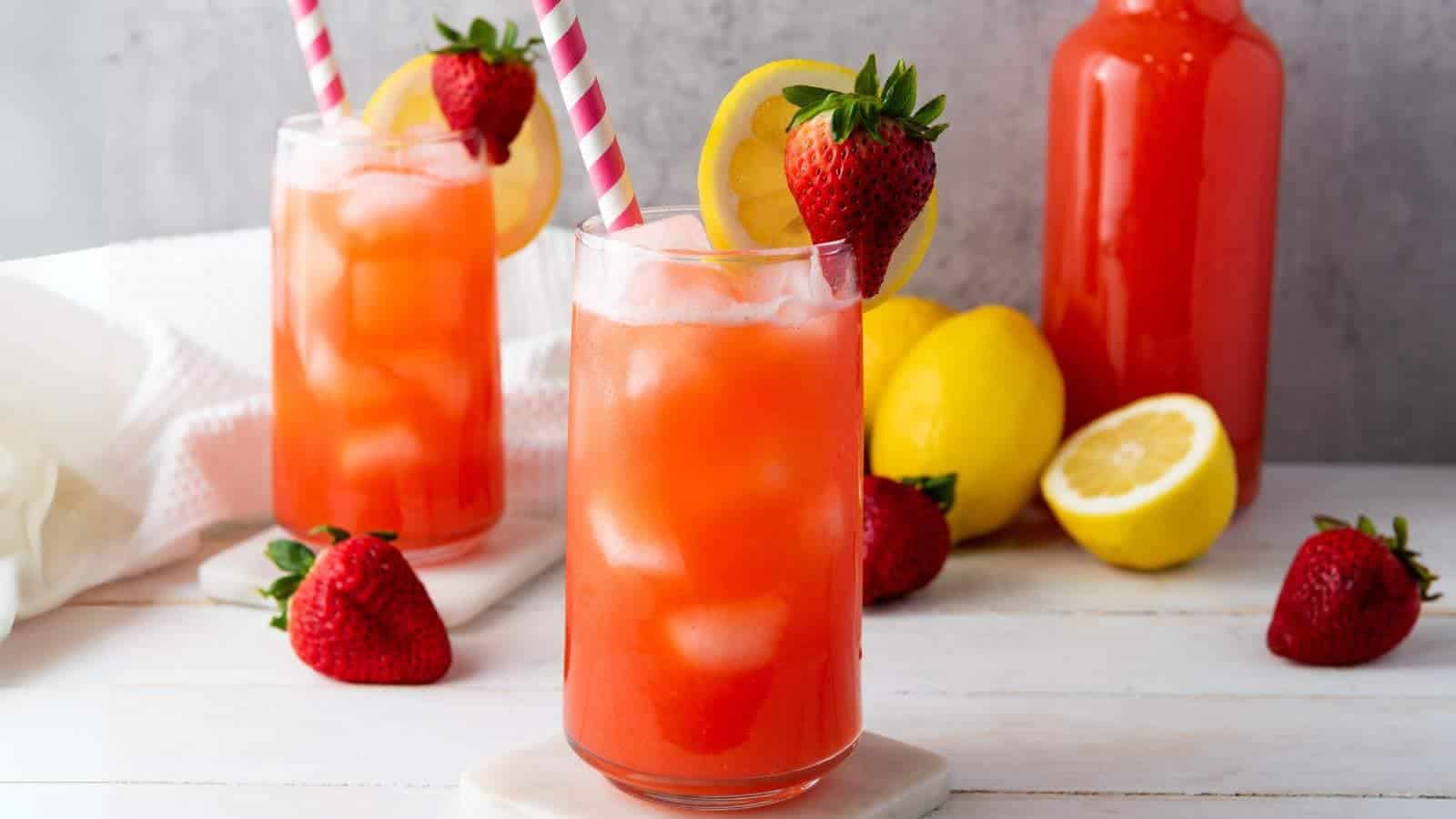 Two glasses of iced strawberry lemonade with pink striped straws, garnished with lemon slices and strawberries. Lemons, strawberries, and a bottle of the drink are in the background.
