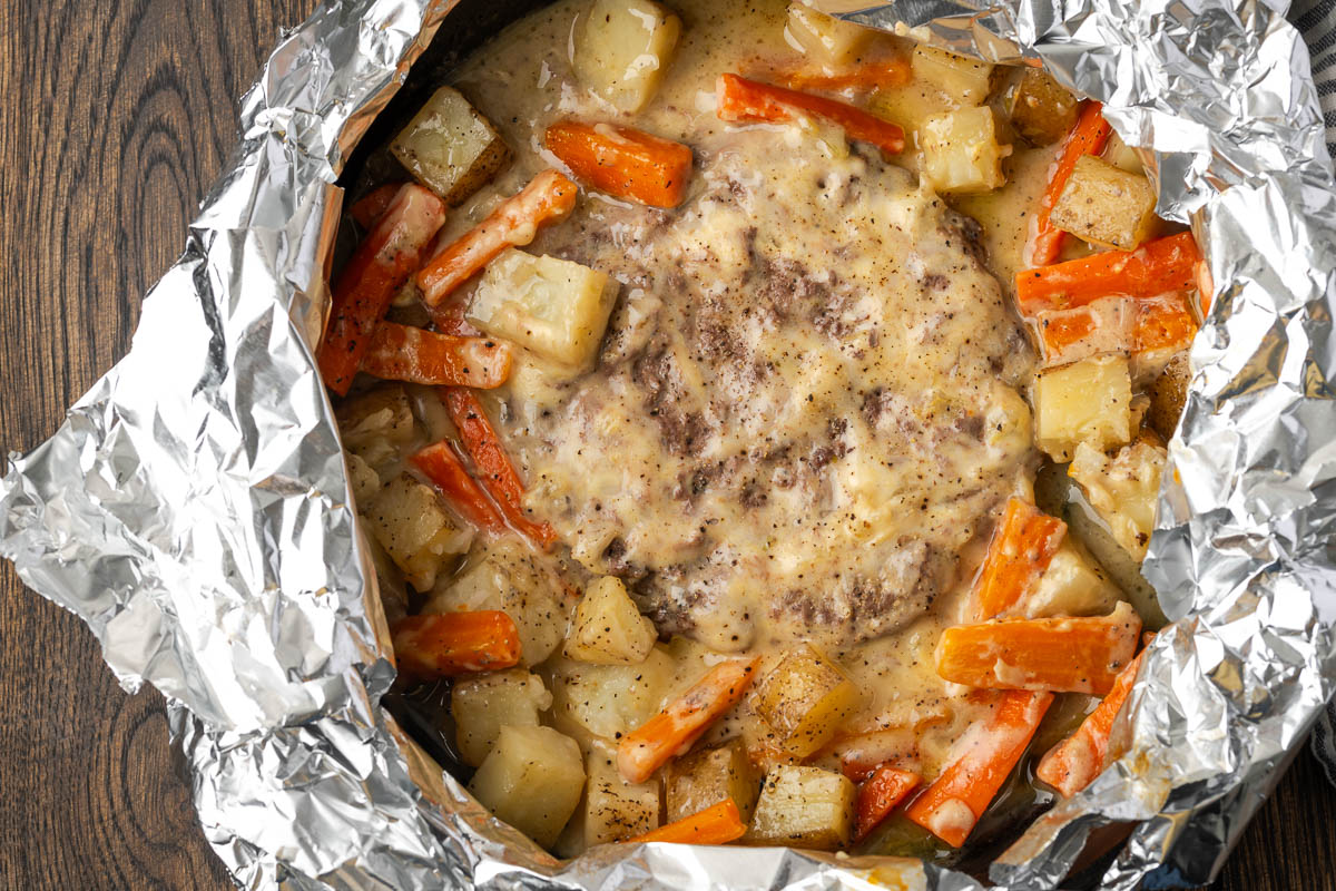 Ground beef patty topped with creamy sauce, surrounded by cooked carrots and potatoes, served in aluminum foil on a wooden surface.