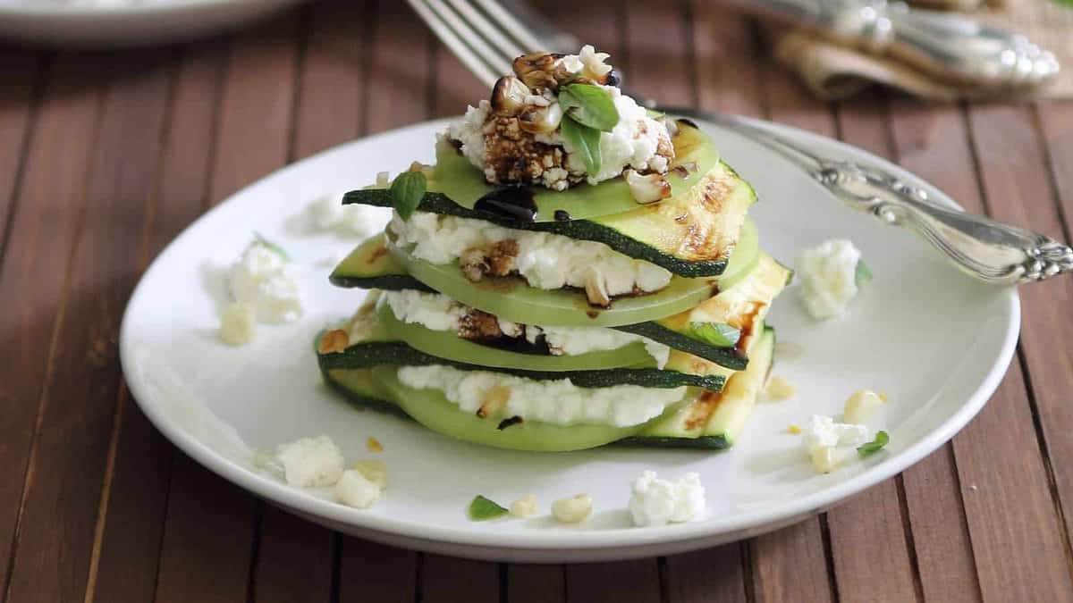 Stacked slices of grilled zucchini layered with ricotta cheese, topped with chopped nuts, herbs, and a drizzle of balsamic glaze on a white plate.