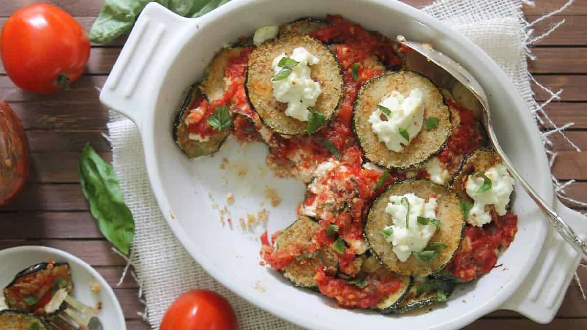 A white baking dish with slices of breaded eggplant topped with tomato sauce, ricotta cheese, and basil. Some portions have been served. Fresh tomatoes and basil are nearby.