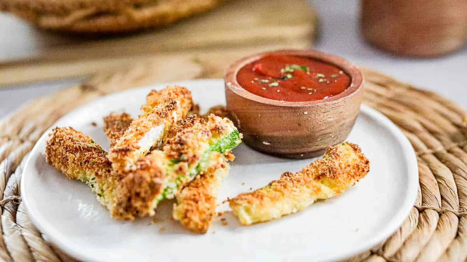 Breaded zucchini sticks are arranged on a white plate next to a small bowl of marinara sauce for dipping, set on a woven placemat.