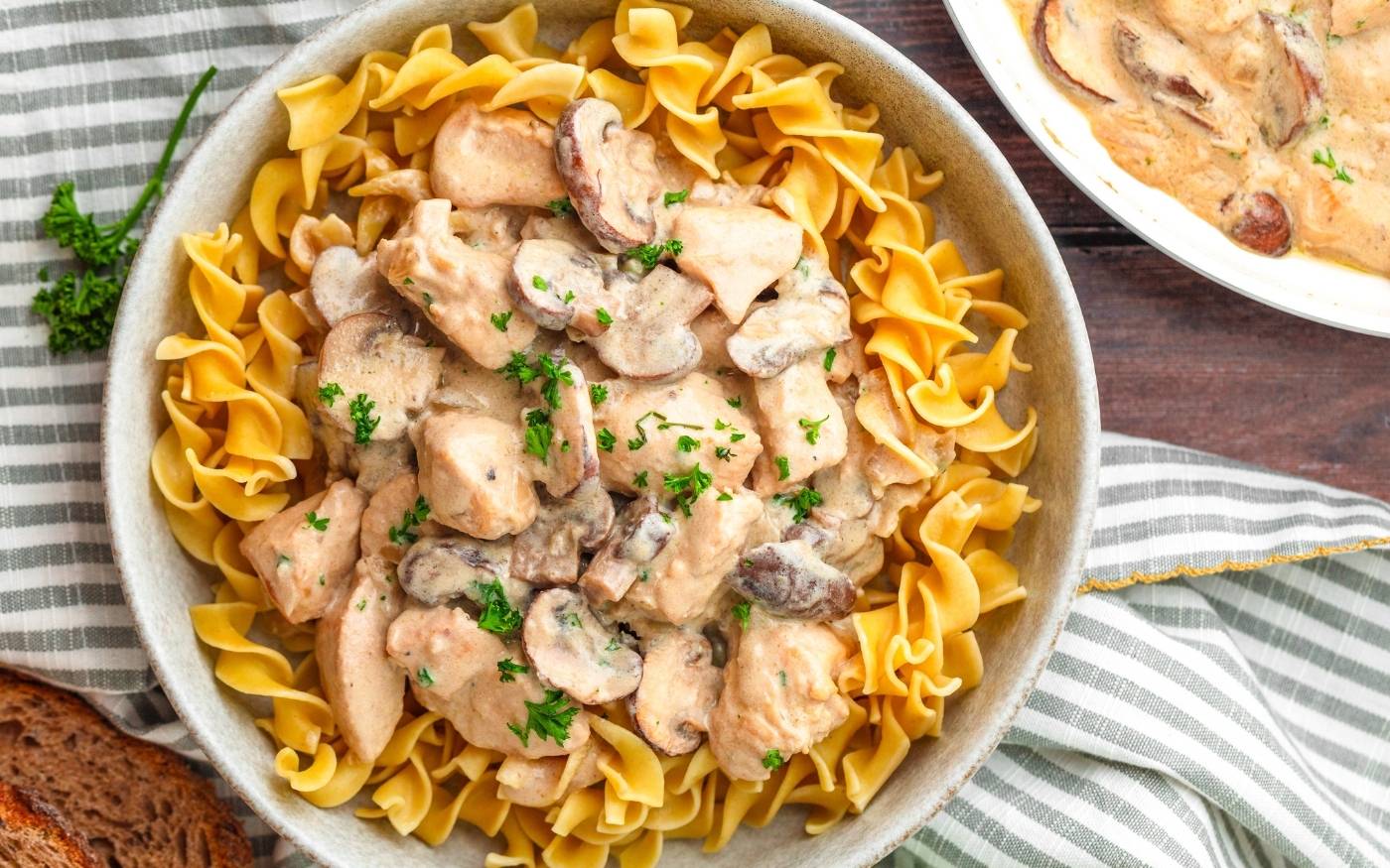 A bowl of egg noodles topped with chicken and mushroom in a creamy sauce, garnished with chopped parsley.