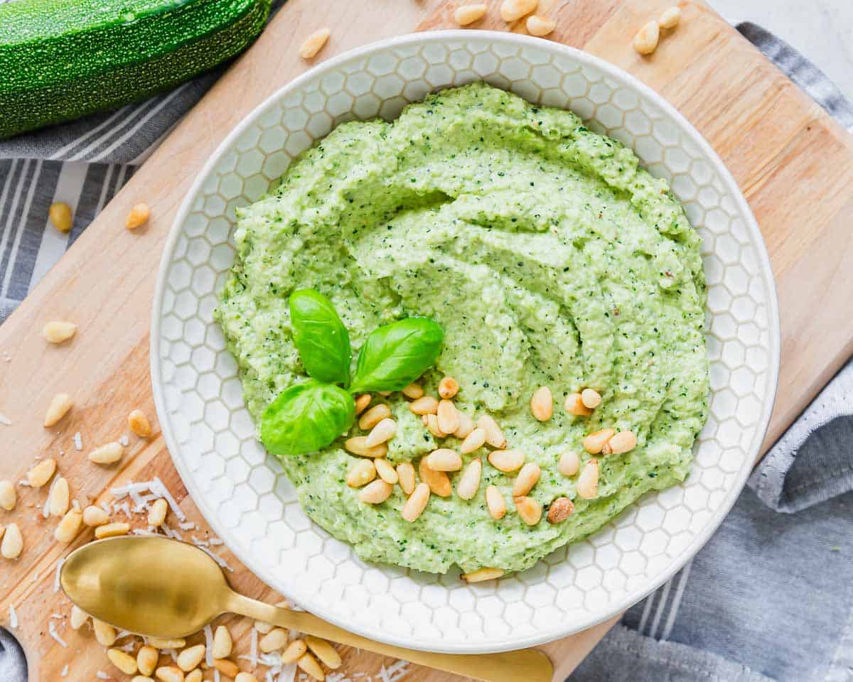 A bowl of green dip garnished with basil leaves and pine nuts, placed on a wooden board with a gold spoon and scattered pine nuts nearby.