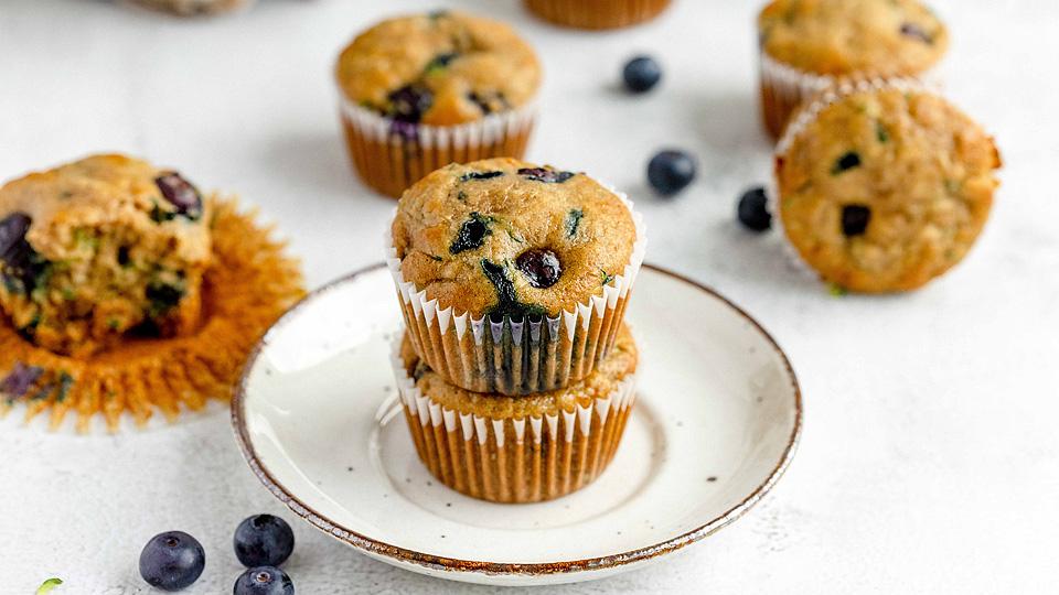 Two blueberry muffins stacked on a white plate, with more muffins and loose blueberries scattered on a white surface in the background.