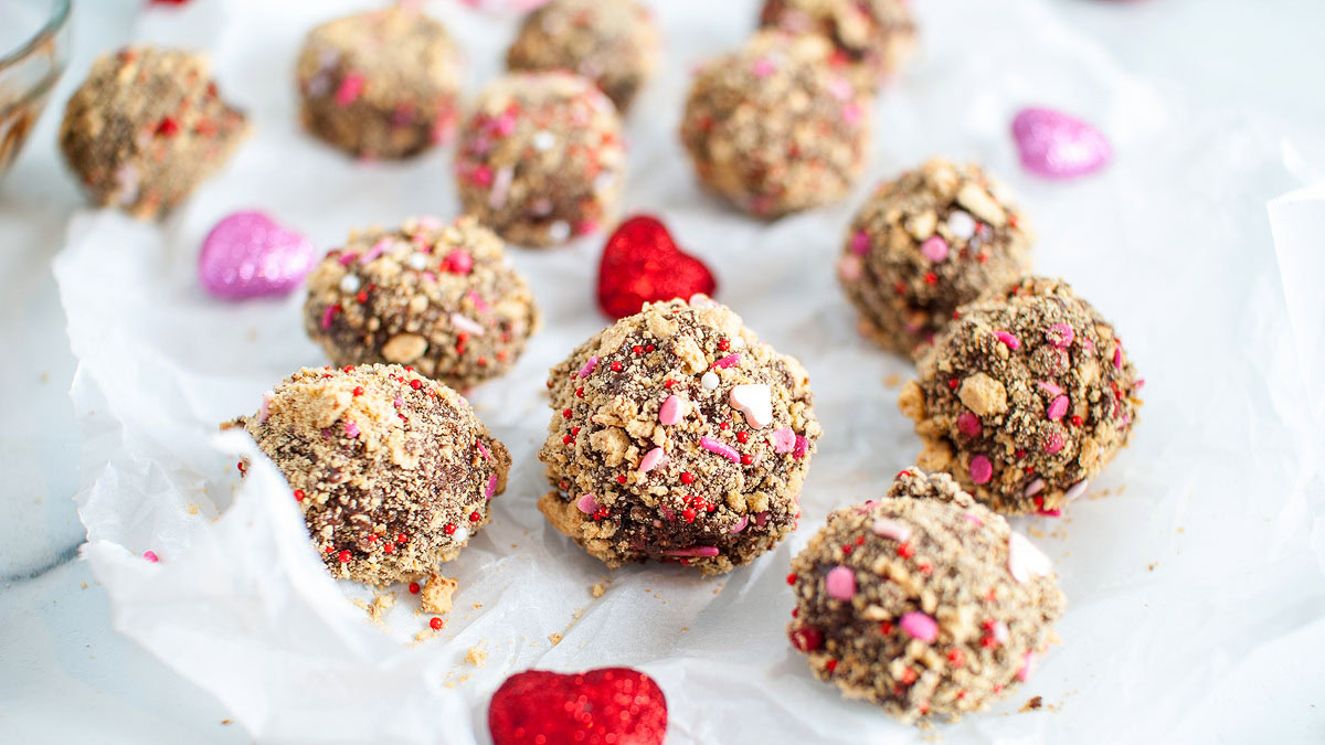 Several chocolate truffles coated in crushed cookies and pink and red sprinkles are arranged on crumpled parchment paper with decorative glitter hearts.