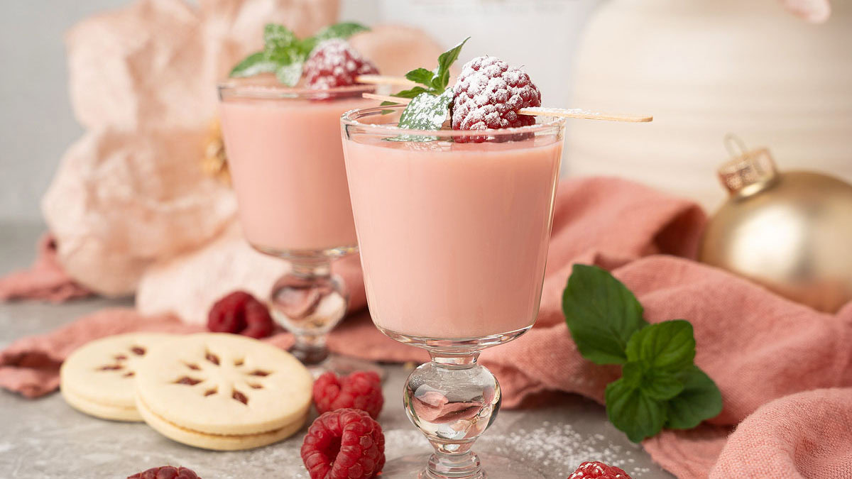 Two glasses of pink mousse dessert topped with raspberries, mint leaves, and powdered sugar, surrounded by cookies and fresh raspberries on a decorated table.