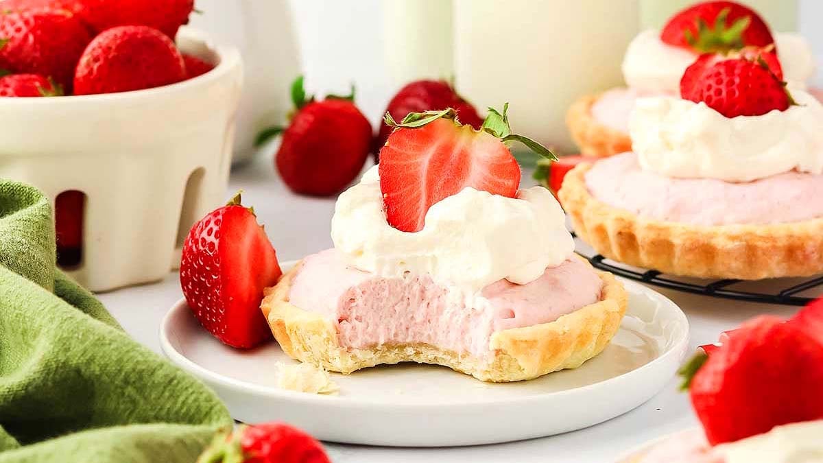 A mini strawberry cream tart with a bite taken out, topped with whipped cream and a strawberry half, sits on a plate with whole strawberries around it.