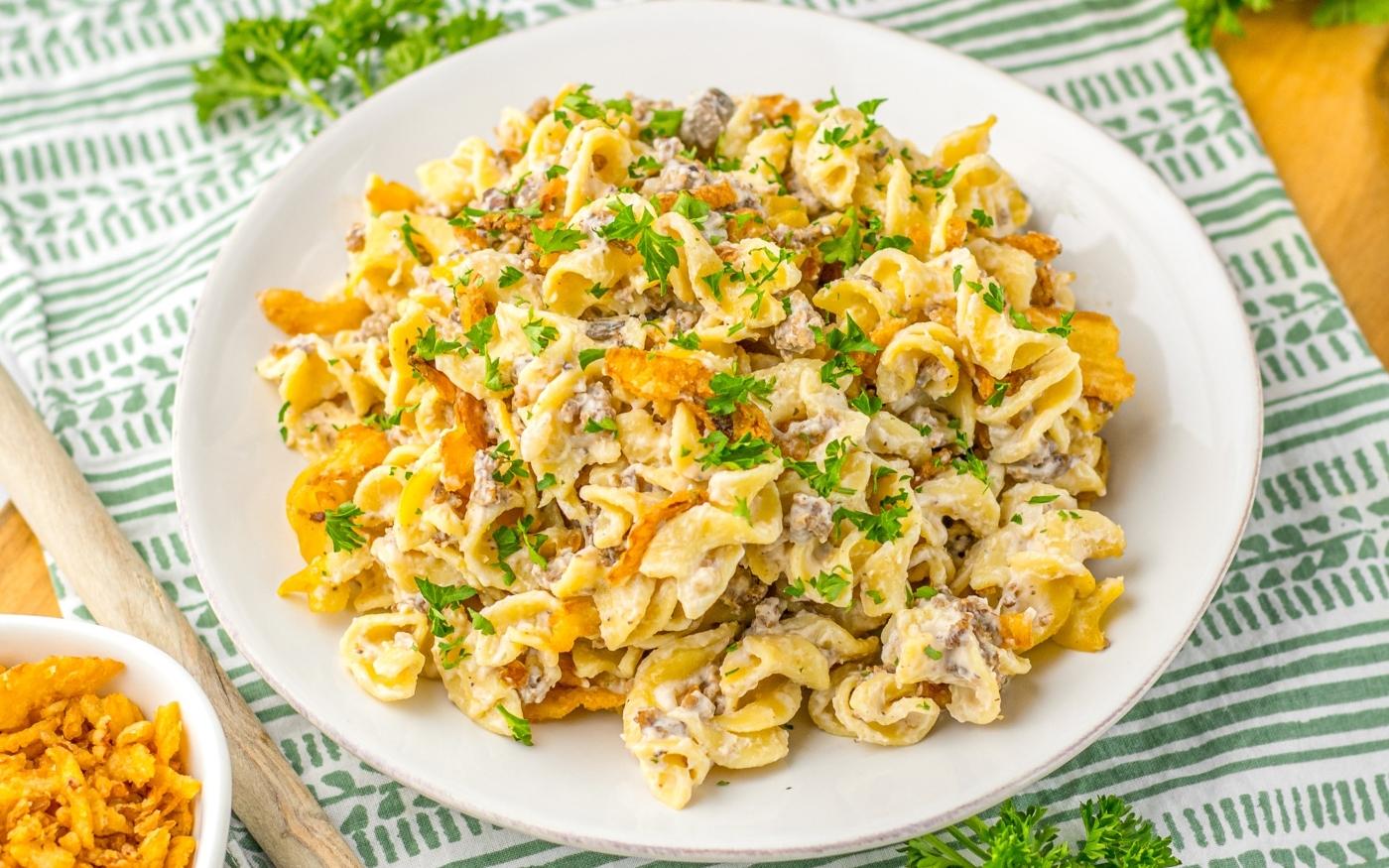 A plate of creamy egg noodles mixed with ground beef, garnished with chopped parsley, served on a white dish on a green and white patterned cloth.