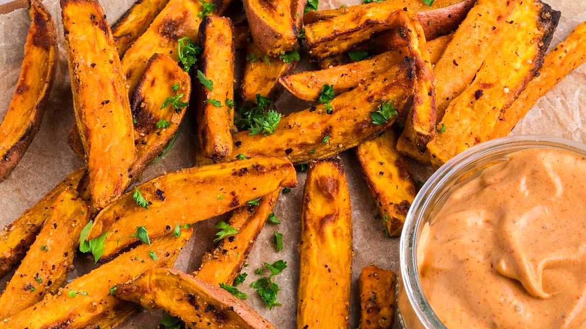 Baked sweet potato fries garnished with chopped herbs next to a bowl of creamy dipping sauce on parchment paper.