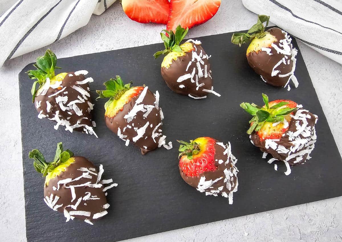 Seven strawberries dipped in chocolate and sprinkled with shredded coconut are arranged on a black slate board, with two strawberry halves and a striped cloth in the background.