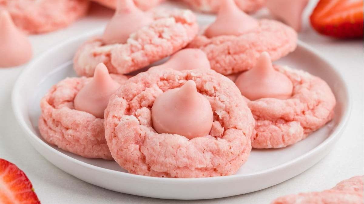 A white plate holds several pink cookies topped with pink chocolate kisses, with a few strawberries visible in the background.