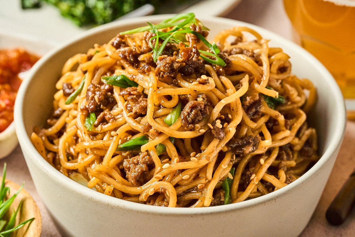 A bowl of stir-fried noodles with ground meat, green onions, and sesame seeds, served on a table.