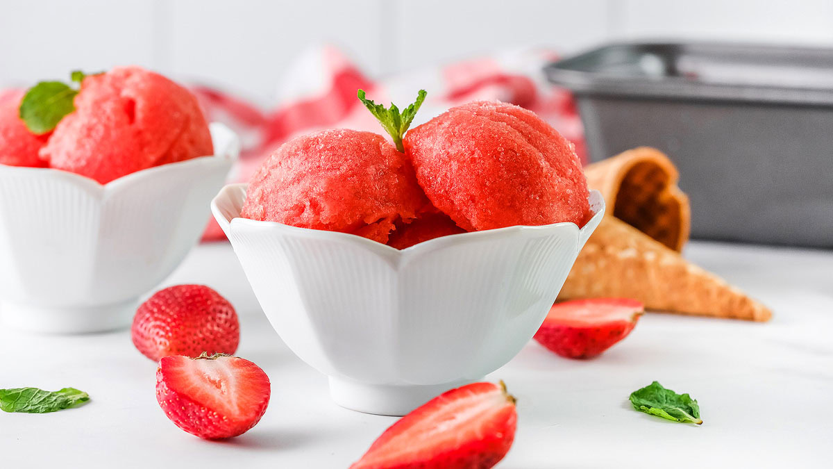 Two scoops of strawberry sorbet in a white bowl, garnished with mint, surrounded by fresh strawberries and an empty waffle cone in the background.