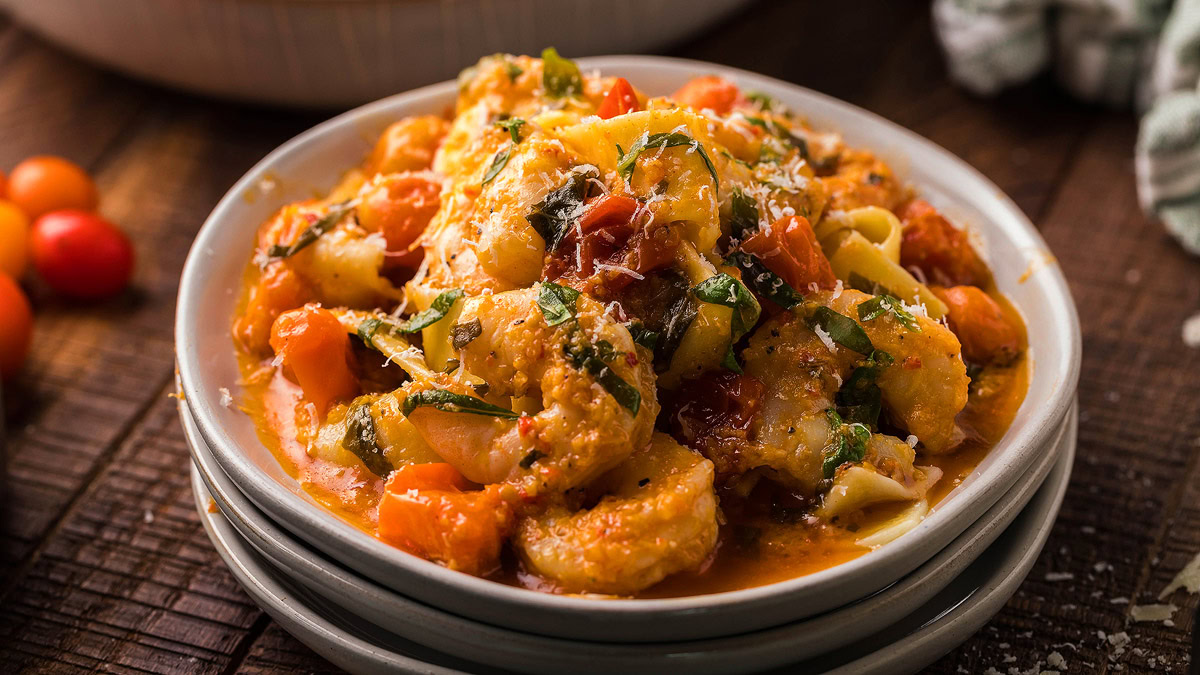 A plate of pasta with tomato sauce, vegetables, herbs, and grated cheese, served on a stack of white plates on a wooden table.