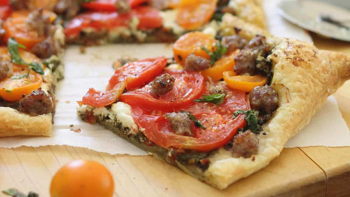 A slice of puff pastry tart topped with sausage, red and yellow tomatoes, fresh herbs, and cheese sits on a wooden board.