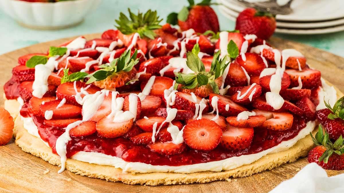 A large strawberry tart with sliced strawberries, a layer of cream, and white icing drizzle sits on a wooden board.