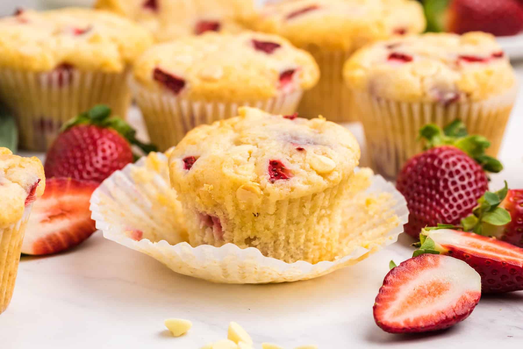A group of strawberry muffins on a white surface, with one muffin unwrapped and fresh strawberries scattered around.
