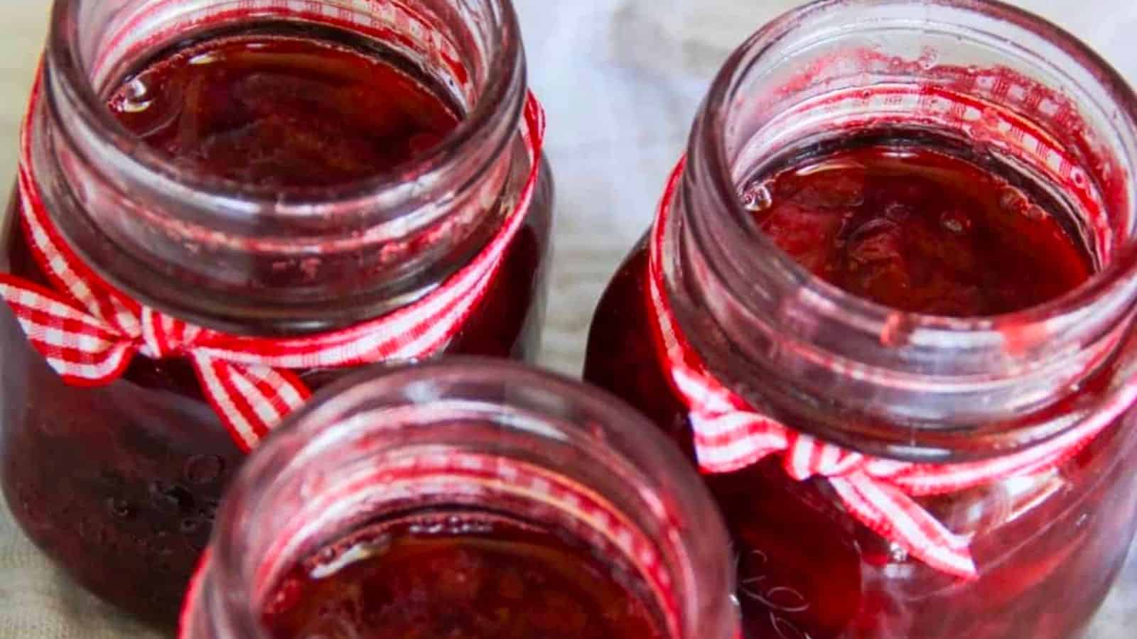 Three glass jars filled with red jam are topped with red and white checkered ribbon bows around their necks, viewed from above.