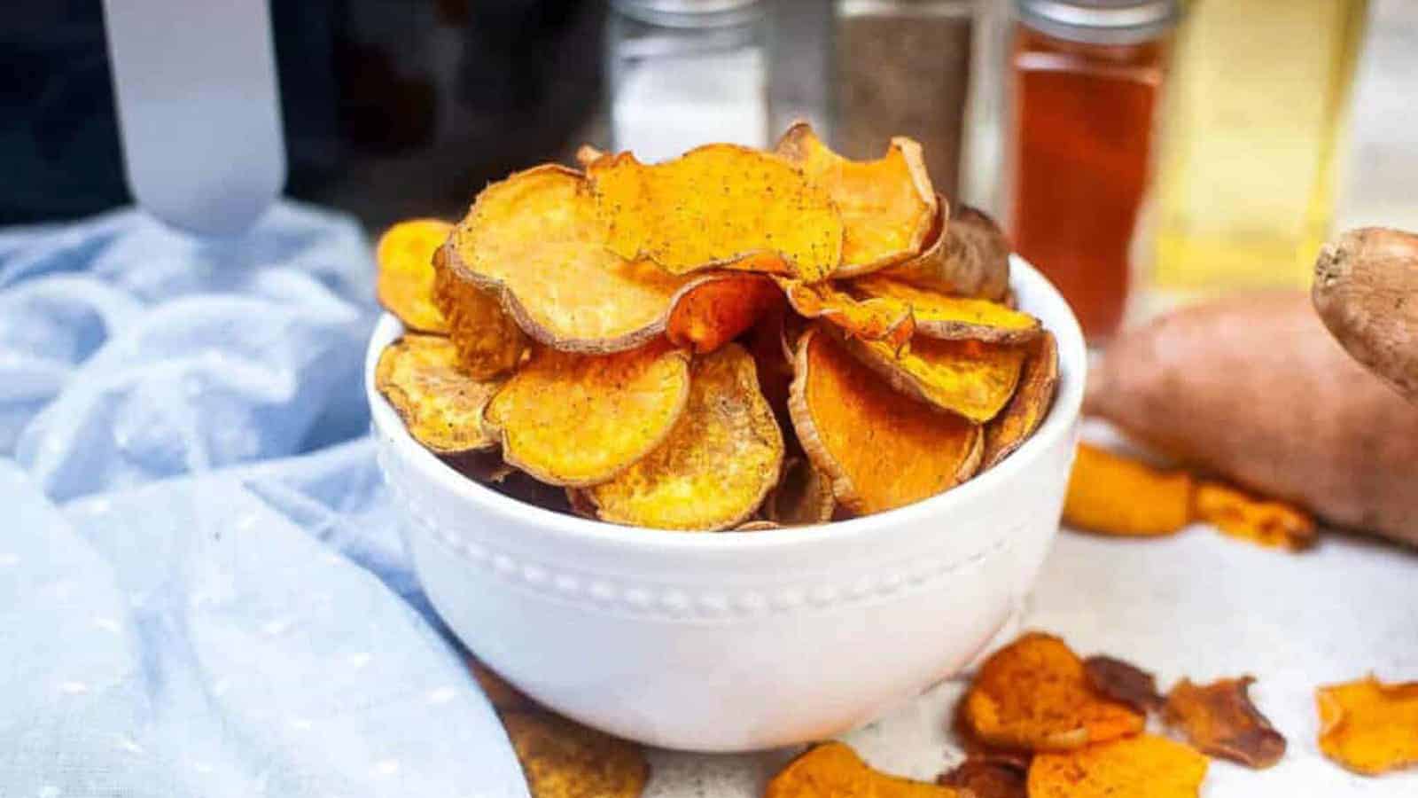 A white bowl filled with crispy, seasoned sweet potato chips sits on a table next to a light blue cloth, spices, and a raw sweet potato.
