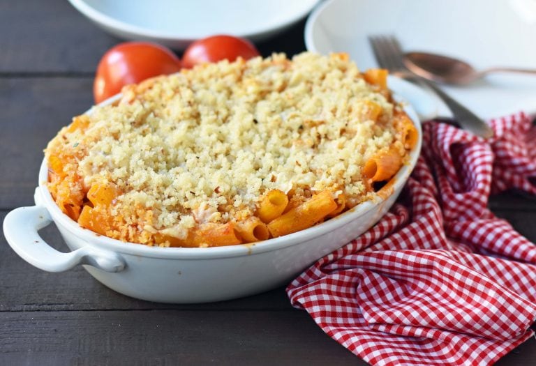 Baked pasta topped with breadcrumbs in a white casserole dish, set on a wooden table with a red checkered cloth, tomatoes, and tableware nearby.