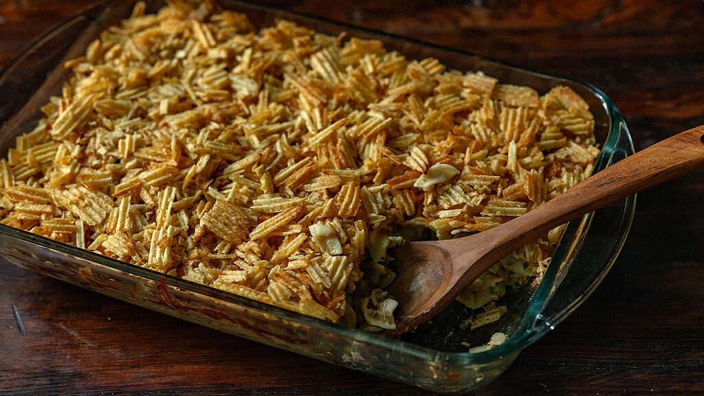 A glass baking dish filled with casserole topped with crumbled potato chips, with a wooden spoon resting inside.