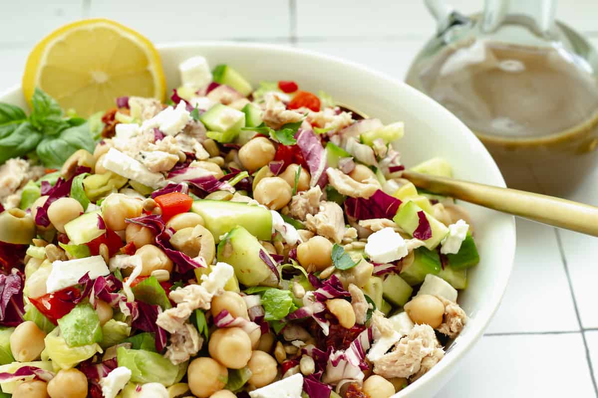 A bowl of chopped salad with chickpeas, feta cheese, cucumber, tuna, and mixed vegetables, garnished with a lemon wedge. A jar of dressing is in the background.