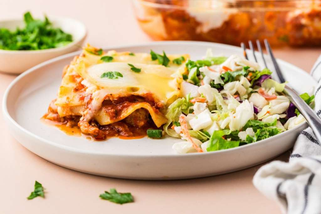 A serving of lasagna topped with melted cheese is plated next to a portion of mixed green salad; a fork and napkin are placed beside the plate.