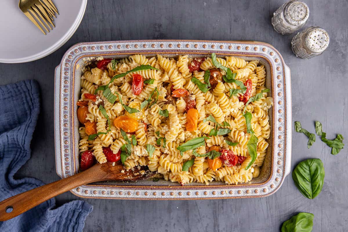 A rectangular dish of rotini pasta with cherry tomatoes, feta cheese, and fresh basil sits on a gray surface next to plates, forks, salt, and pepper shakers.