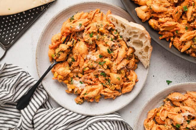 A plate of pasta with tomato sauce, vegetables, and ground meat, garnished with parsley and served with a piece of bread. A black fork rests on the plate.