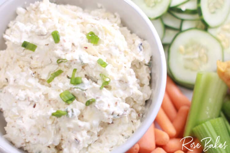A bowl of creamy dip garnished with chopped green onions, surrounded by sliced cucumbers, celery sticks, and baby carrots.