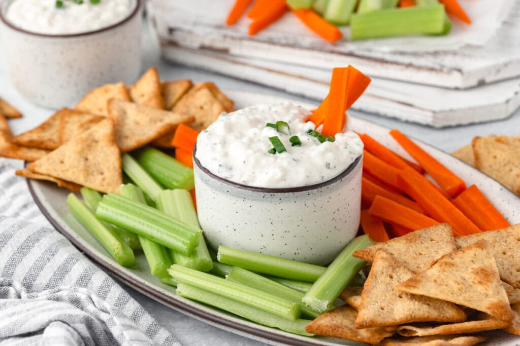 A platter with a bowl of cottage cheese dip, surrounded by celery sticks, carrot sticks, and pita chips.