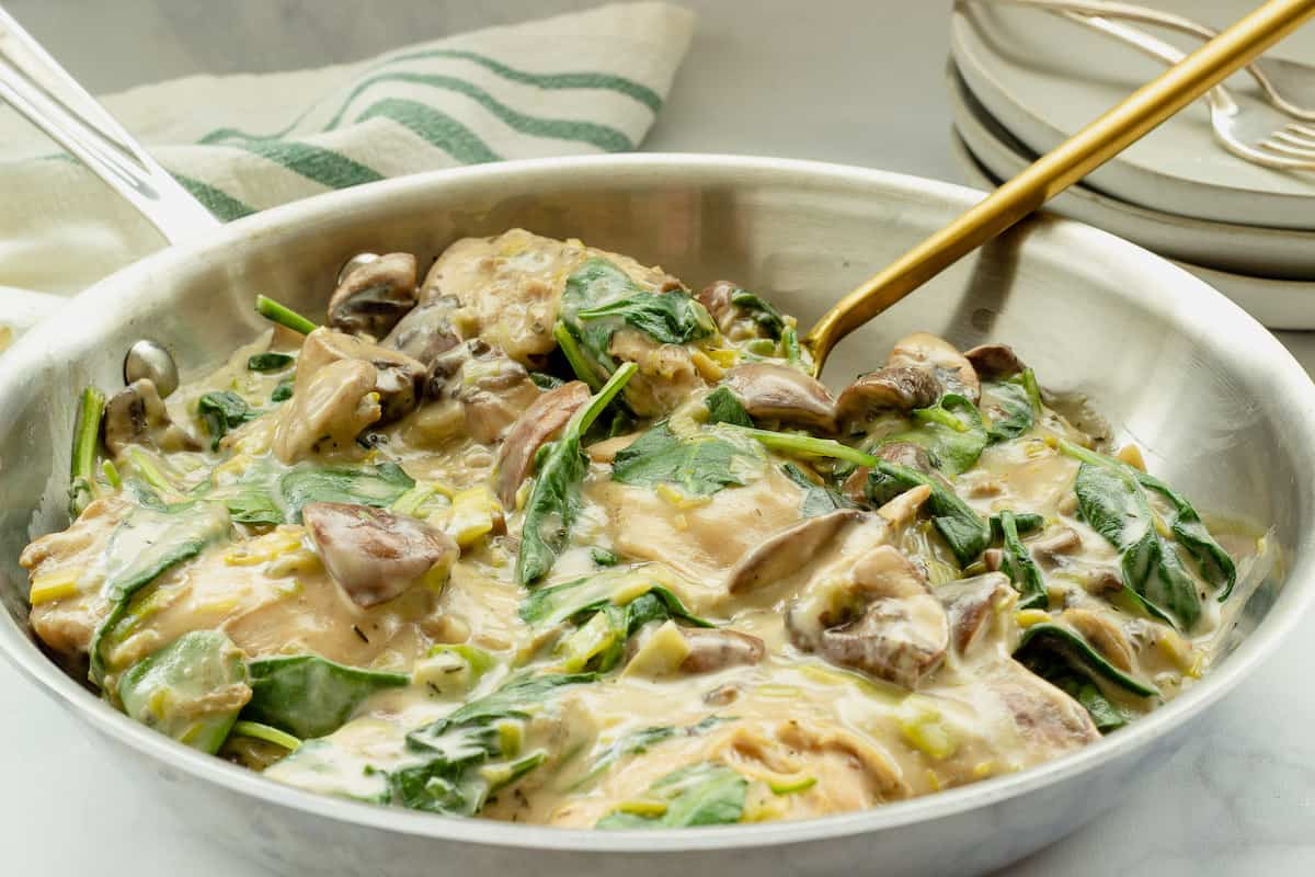 A skillet filled with creamy chicken, mushrooms, and spinach, with a gold spoon beside it and a striped towel in the background.
