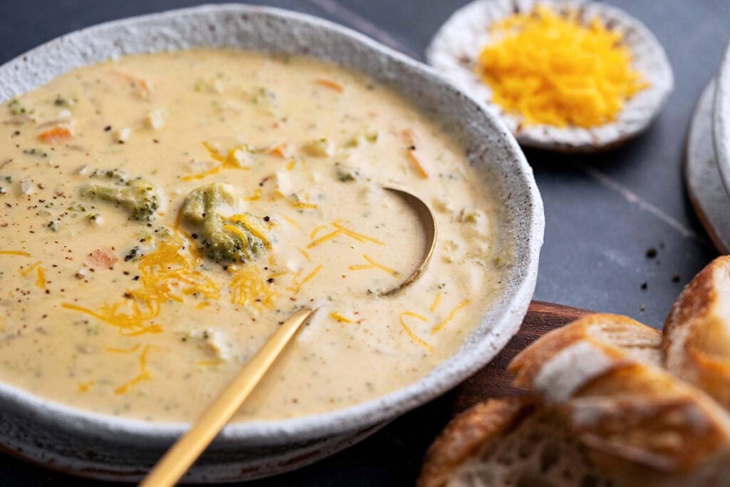 A bowl of creamy broccoli cheddar soup with a spoon, topped with shredded cheese, next to slices of bread and a small plate of grated cheddar.