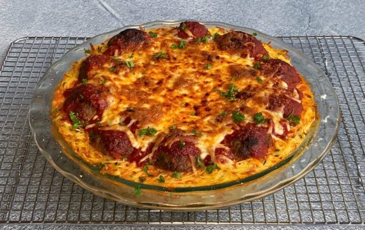 A baked spaghetti pie topped with marinara sauce, browned meatballs, melted cheese, and parsley sits on a cooling rack.