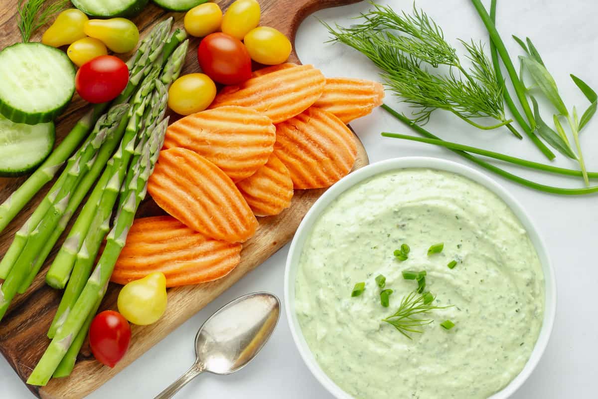 A platter with sliced cucumbers, cherry tomatoes, asparagus, and crinkle-cut carrots next to a bowl of green herb dip, garnished with fresh dill and chives.