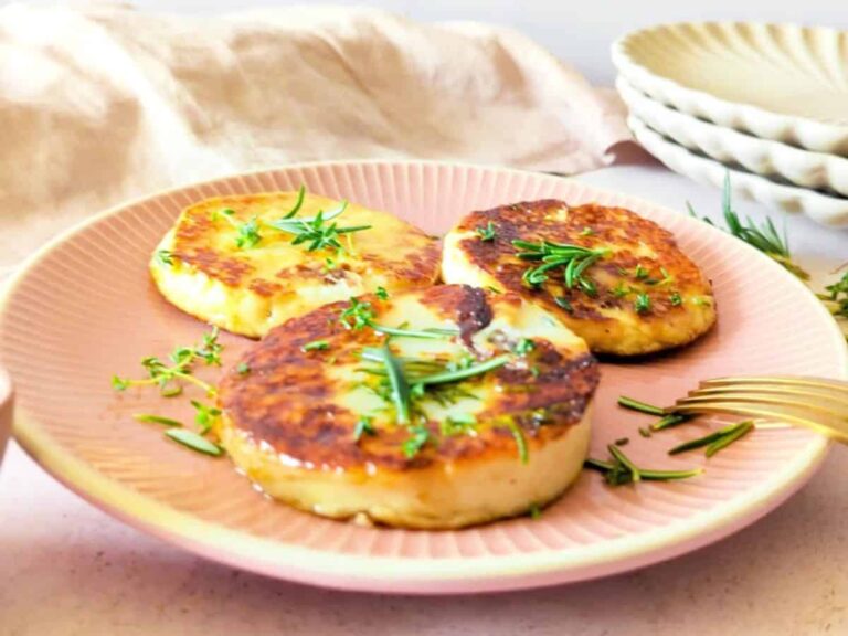 Three golden-brown fried cheese rounds garnished with fresh herbs are served on a pink plate next to a gold fork.