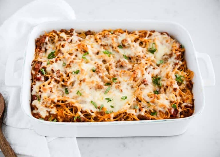 A white baking dish filled with baked spaghetti topped with melted cheese and sprinkled with chopped herbs, sitting on a white surface.