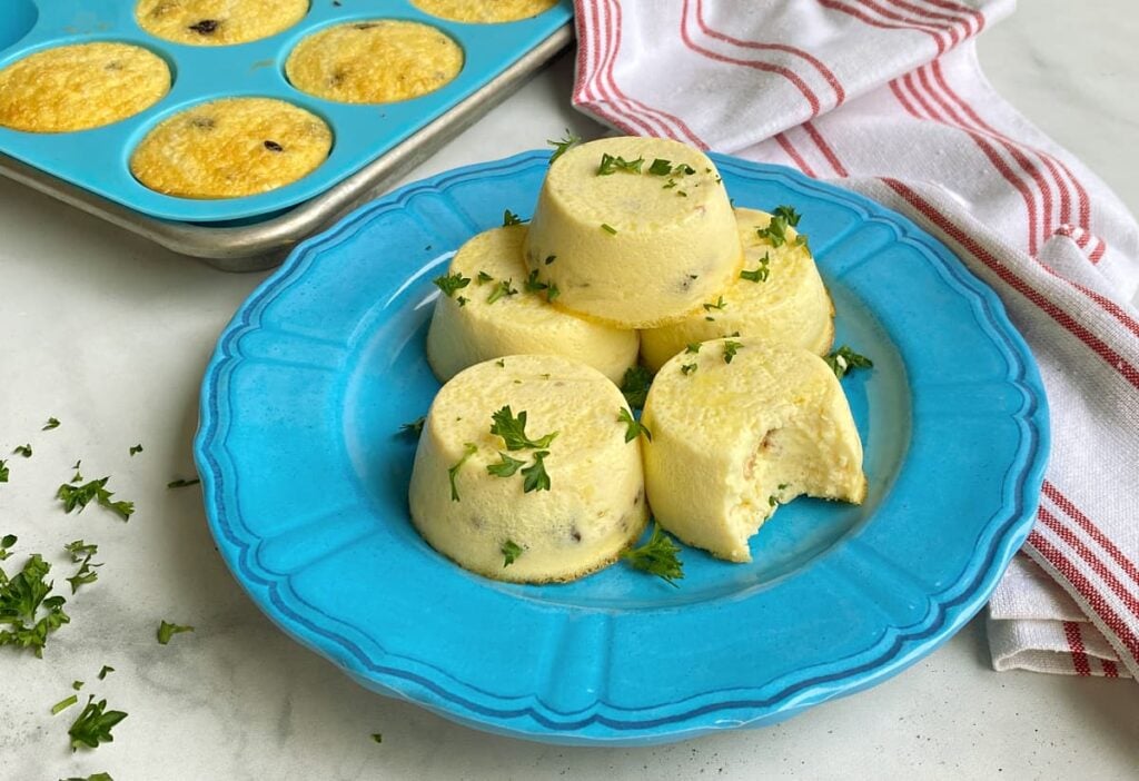 Four egg bites garnished with chopped herbs are stacked on a blue plate, with a bite taken out of one. A muffin tin with more egg bites and a striped towel are in the background.
