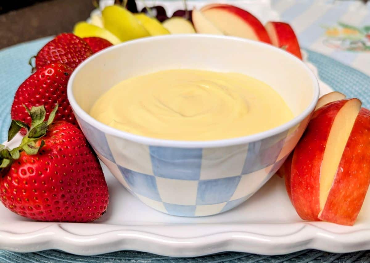 A bowl of creamy dip is surrounded by fresh strawberries, apple slices, and grapes on a white plate.