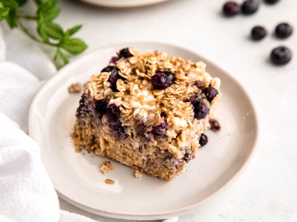 A square slice of baked oatmeal with blueberries on a white plate, with some fresh blueberries and a sprig of mint nearby.