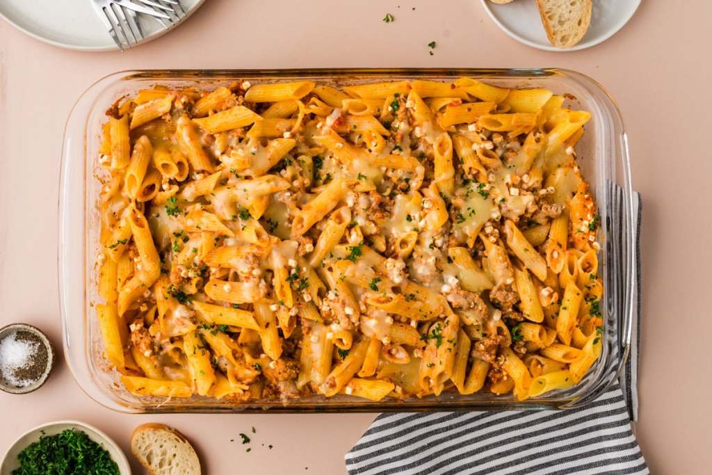 A glass baking dish filled with baked penne pasta topped with melted cheese and herbs, placed on a light surface next to bread slices and a striped napkin.