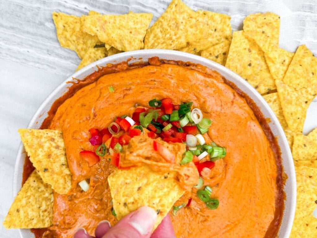 A hand dips a tortilla chip into a bowl of orange cheese dip topped with chopped green onions and red peppers, surrounded by more tortilla chips.