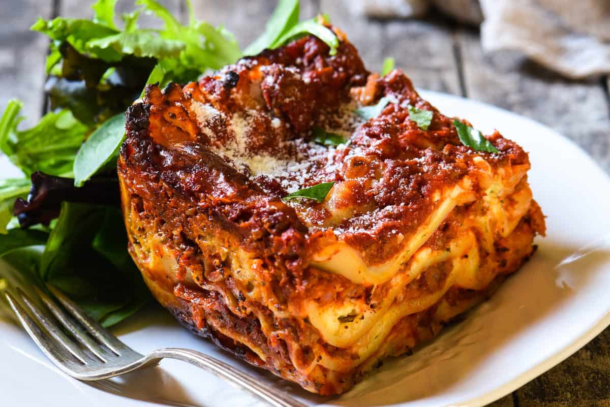 A slice of lasagna with layers of pasta, meat sauce, and cheese on a white plate, served with a side of mixed greens and a fork.