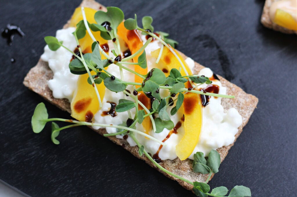 A crispbread topped with cottage cheese, yellow bell pepper slices, microgreens, and a drizzle of balsamic glaze on a dark surface.