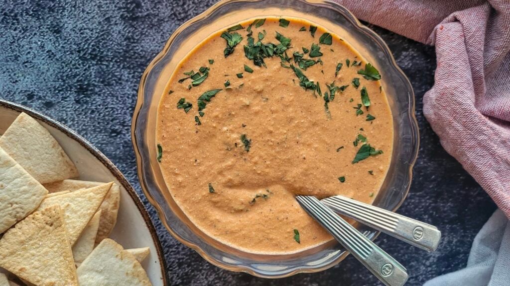 A bowl of creamy orange dip garnished with chopped herbs, with two spoons inside, next to a plate of pita chips on a dark textured surface.