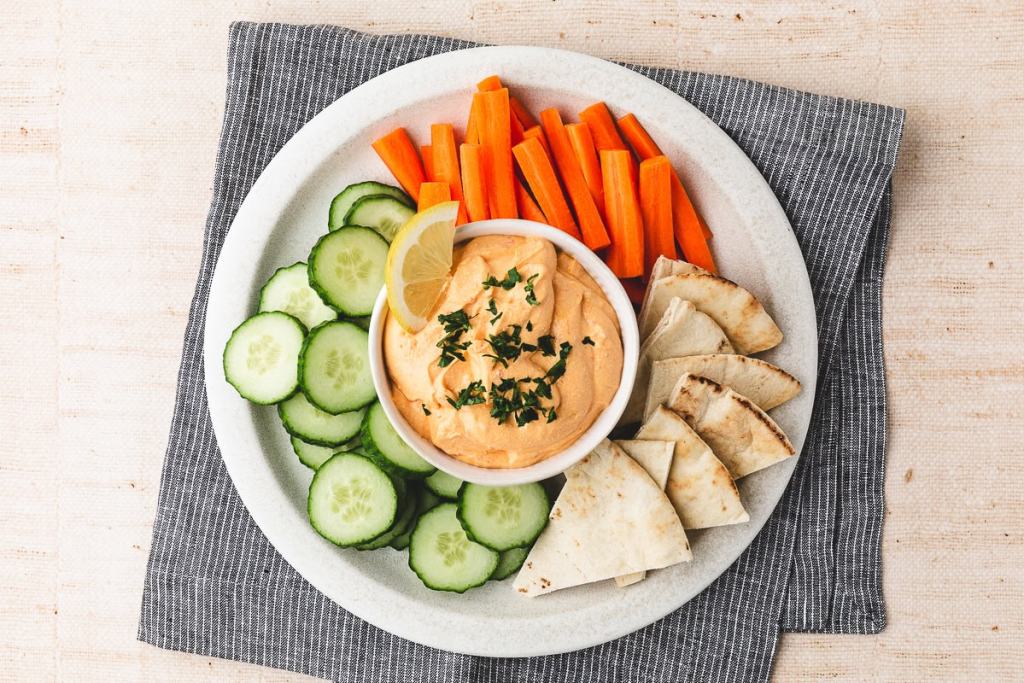 A plate with sliced cucumbers, carrot sticks, pita wedges, and a bowl of hummus topped with herbs and a lemon slice.