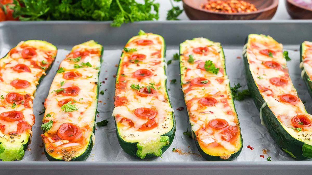 Six zucchini boats filled with melted cheese and sliced cherry tomatoes are arranged in a row on a baking sheet, topped with herbs and placed on parchment paper.